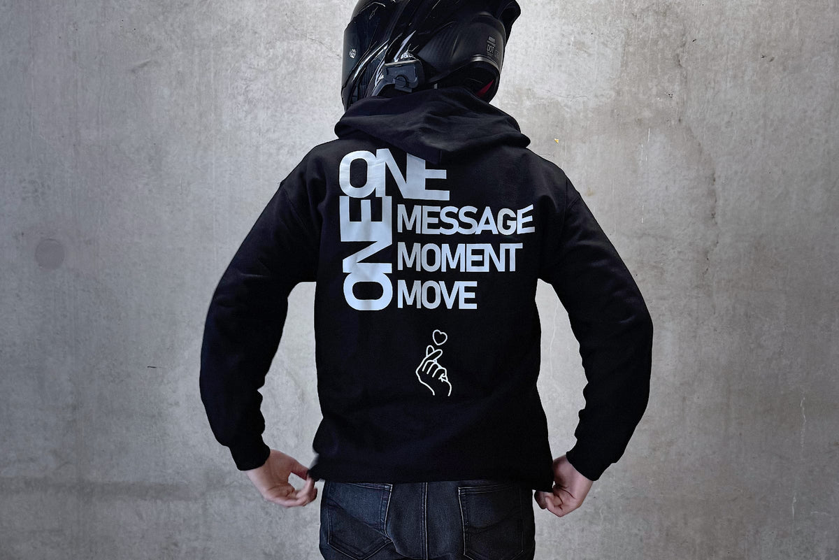 A person standing with their back to the camera, wearing a black hoodie with bold white text on the back reading 'ONE MESSAGE MOMENT MOVE', dark jeans, white sneakers, and a black motorcycle helmet against a gray concrete wall.