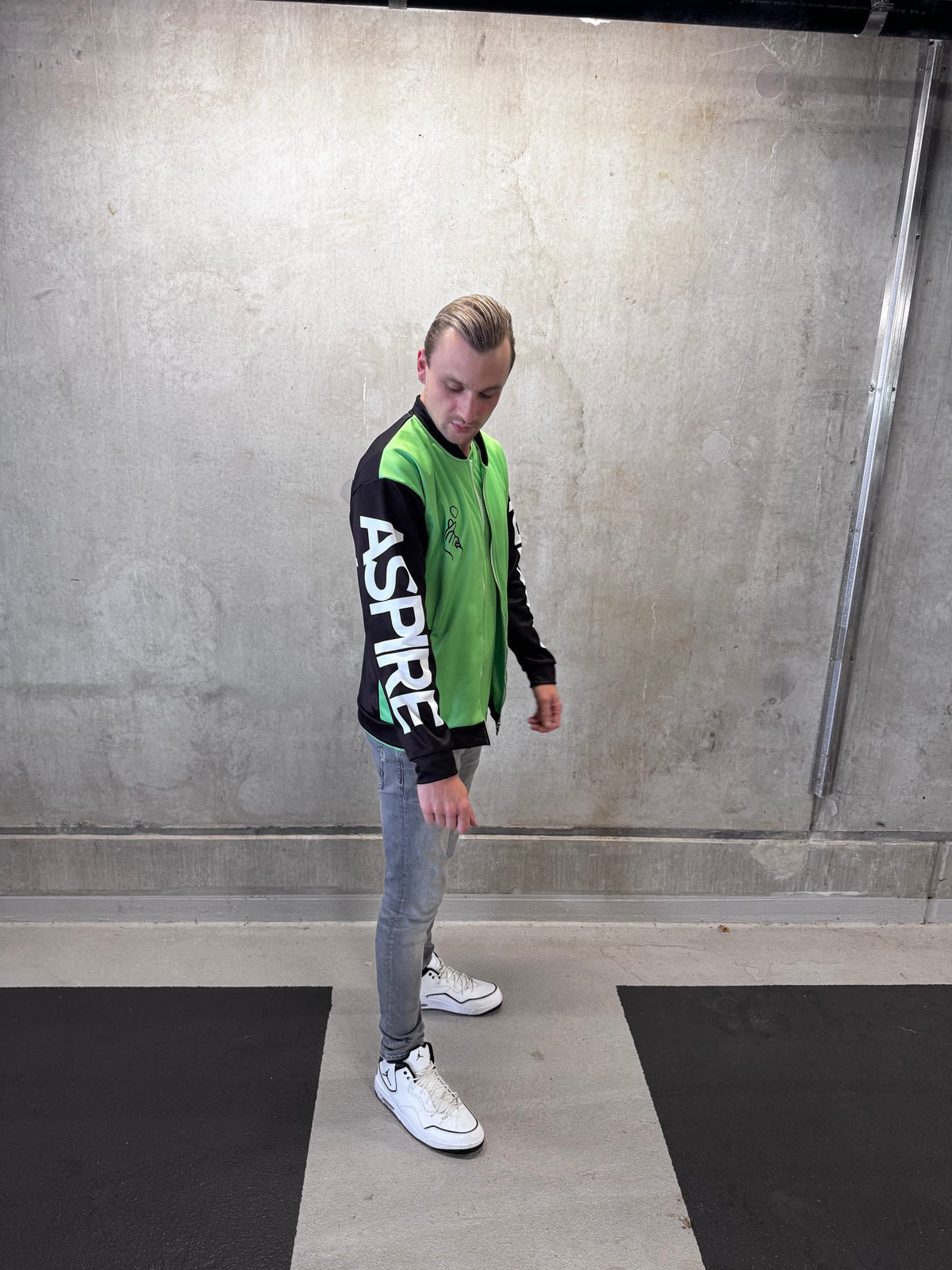 A young man wearing a green and black AZPR summer jacket featuring ASPIRE written on the right arm, gray jeans, and white sneakers, standing in a concrete-walled indoor space.